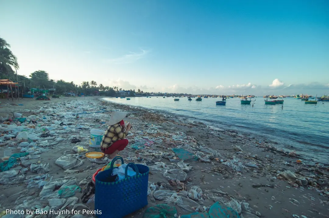 Plastic waste along a shoreline with a person and fishing boats in the background