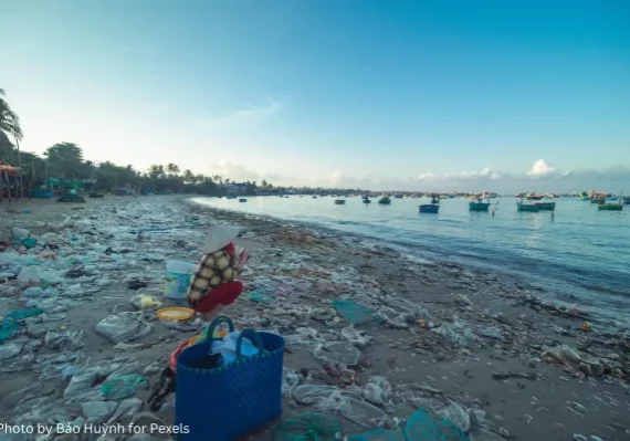 Plastic waste along a shoreline with a person and fishing boats in the background